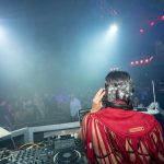 DJ se apresenta diante de um público animado em uma casa noturna, sob iluminação colorida.