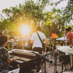 Músicos se apresentam em um palco ao ar livre durante o pôr do sol, diante do público.
