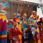 Artistas circenses fantasiados, em pernas de pau, entretêm o público durante um evento ao ar livre.