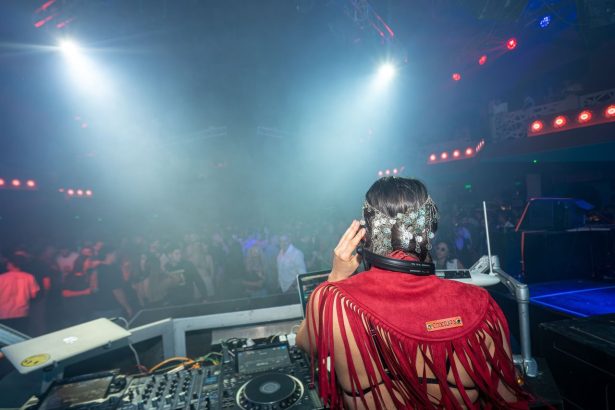 DJ se apresenta diante de um público animado em uma casa noturna, sob iluminação colorida.