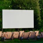 Espaço de cinema ao ar livre, com cadeiras listradas diante de uma tela em branco, cercado por vegetação.