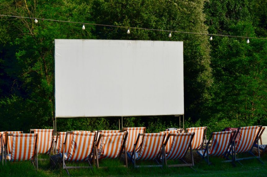 Espaço de cinema ao ar livre, com cadeiras listradas diante de uma tela em branco, cercado por vegetação.